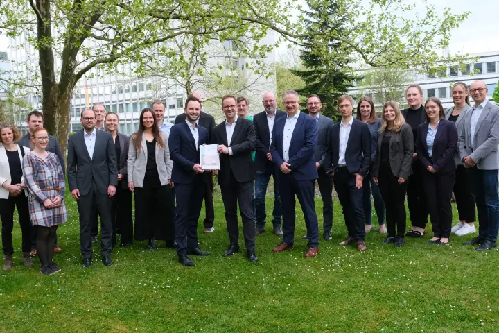 Eine Gruppe von mehreren Personen, die meisten in formeller oder legerer Geschäftskleidung, posiert für ein Foto im Freien auf einer Grasfläche vor einem Bürogebäude.