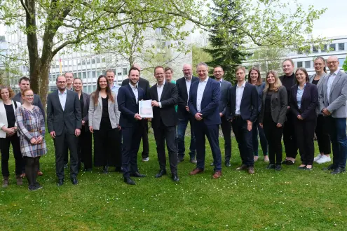 Eine Gruppe von mehreren Personen, die meisten in formeller oder legerer Geschäftskleidung, posiert für ein Foto im Freien auf einer Grasfläche vor einem Bürogebäude.