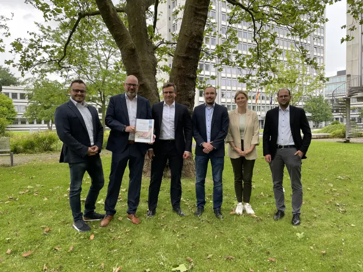 Sechs Personen, fünf Männer und eine Frau, stehen im Freien vor einem Bürogebäude. Die Gruppe trägt formelle bis legere Geschäftskleidung. Ein Mann in der Mitte hält ein gerahmtes Dokument oder eine Urkunde in die Kamera.
Die Personengruppe steht auf einer Rasenfläche. Im Hintergrund befindet sich ein modernes Bürogebäude mit vielen Fenstern und ein großer Baum mit grünen Blättern.