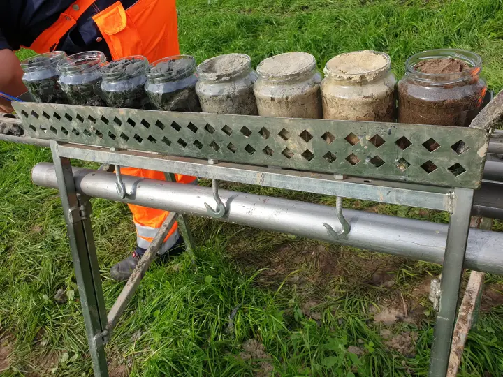 Auf dem Bild sind Bodenproben zu sehen, die im Rahmen von Baugrunduntersuchungen für ein großes Infrastrukturprojekt, wie die Amprion-Erdkabeltrasse A-Nord, entnommen wurden.
Beschreibung
Eine Reihe von sieben Glasgefäßen, gefüllt mit Erde unterschiedlicher Farbe und Konsistenz, steht in einem speziell angefertigten Metallgestell.
Bodenproben: Die Proben reichen von sehr dunkler, feuchter Erde links, die vermutlich Oberboden darstellt, bis zu hellerem, sandigem oder lehmigem Material rechts. Die Gläser links haben Metallbügelverschlüsse, während die rechten Gläser offen sind oder lose aufliegende Deckel haben.
Ausrüstung: Das grüne Metallgestell mit rautenförmigen Aussparungen dient der sicheren Aufbewahrung und dem Transport der Proben. Darunter ist eine silberne Metallstange mit Haken zur Befestigung von Werkzeugen oder weiterem Equipment angebracht.
Person und Umgebung: Im Hintergrund ist eine Person in orangefarbener Warnschutzkleidung (Bundhose) und blauem Oberteil zu sehen, die die Proben entnimmt oder arrangiert. Der Untergrund ist eine grüne Wiese mit einigen nackten Erdflecken.