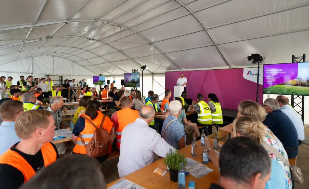 Präsentation: Im Hintergrund steht ein Mann im weißen Hemd an einem Rednerpult und spricht zu den Anwesenden. Flankiert wird er von zwei großen Bildschirmen.
Der linke Bildschirm zeigt das Amprion-Logo auf einem pinkfarbenen Hintergrund.
Der rechte Bildschirm zeigt eine Grafik oder ein Foto, das den Bau eines Projekts in einer ländlichen Umgebung mit markanten Strommasten im Hintergrund darstellt.