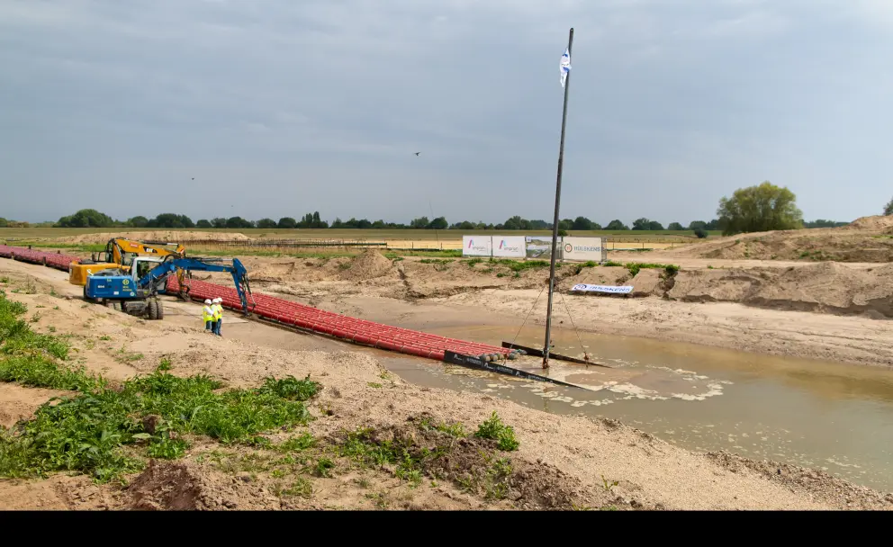 Vordergrund: Im Vordergrund befindet sich eine Aufschüttung aus hellem Sand und Kies. Zwei Bauarbeiter in orangefarbener Warnschutzkleidung und Schutzhelmen stehen in der Nähe eines blauen Kettenbaggers.
Ausrüstung: Ein langes, rotes Bündel aus Kabelschutzrohren, die durch Auftriebskörper zusammengehalten werden, ragt über das Ufer in das braune Wasser. Diese Rohre wurden für das Einziehen in eine unterirdische Bohrung vorbereitet, um den Rhein zu unterqueren. Eine vertikale Stange mit einer Flagge ist an den Rohren befestigt, um deren Position im Wasser zu markieren.
Hintergrund: Hinter der Baustelle erstreckt sich eine grüne Landschaft mit Bäumen und Büschen am Horizont. Weiße Banner, auf denen unter anderem "AMPRION" und "A-NORD" zu lesen sind, sind am oberen Rand der Baugrube angebracht. Der Himmel ist bewölkt. 