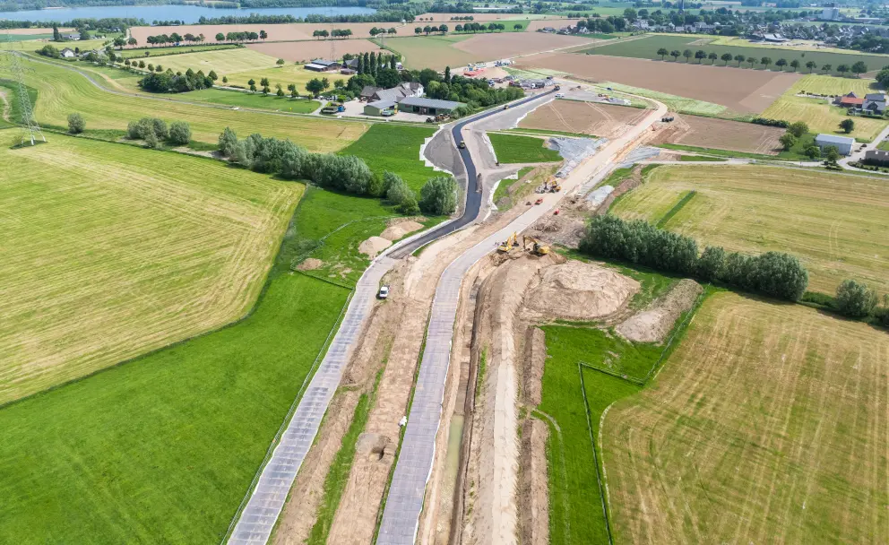 Vordergrund und Aktivitäten: Im Vordergrund verläuft die Baustelle diagonal durch grüne Felder. Es sind mehrere Fahrspuren zu erkennen, teilweise asphaltiert oder mit Schotter und Vlies ausgelegt. Im zentralen Bereich der Baustelle sind kleinere Baumaschinen wie Bagger und Fahrzeuge erkennbar.
Umgebung: Die Baustelle schneidet durch landwirtschaftlich genutzte Flächen. Im oberen linken Teil des Bildes ist ein großes Gewässer oder ein See sichtbar, der an die Felder grenzt. Mehrere Gebäude, die zu einem landwirtschaftlichen Betrieb oder einem kleinen Gewerbegebiet gehören, stehen in der Mitte des Bildes in der Nähe der Trasse. Die Landschaft ist von Bäumen, Büschen und Wiesen geprägt. Im oberen rechten Bereich sind weitere Ackerflächen und Wohnhäuser zu sehen.
Atmosphäre: Der Himmel ist blau mit leichten Wolken, was auf helles Tageslicht hindeutet.