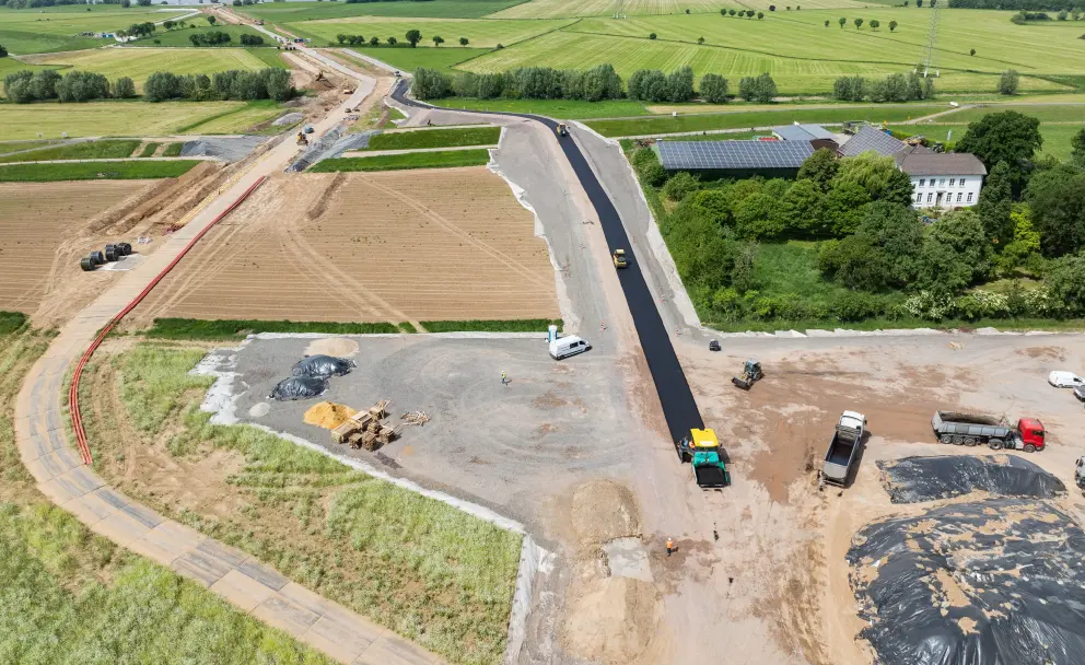 Baustellentrasse: Die Baustelle verläuft als breiter Streifen durch landwirtschaftliche Felder. Im linken Teil sind braune, offene Gräben und vorbereitete Flächen sichtbar, die durch orange und rote Markierungen abgegrenzt sind.
Asphaltierungsarbeiten: Im rechten Teil wird aktiv am Straßenbau gearbeitet. Ein gelber Asphaltfertiger trägt eine schwarze Asphaltschicht auf den vorbereiteten Schotteruntergrund auf. Ein schwarzer LKW mit Kippmulde steht bereit.
Gebäude und Umgebung: Im mittleren rechten Bildbereich befindet sich ein weißes Gebäude mit einem roten Dach, umgeben von Bäumen. Dahinter liegen weitere grüne Felder. Im Hintergrund, am Horizont, ist ein großes Gewässer erkennbar, was auf die Nähe zum Rhein hindeutet.
Hintergrund: Die Landschaft besteht aus weiten Feldern, Wiesen und kleinen Wäldchen, unter einem hellblauen Himmel mit wenigen Wolken. 