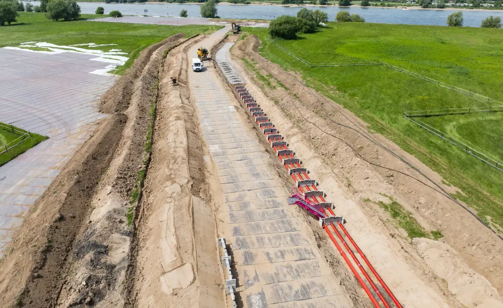 Baustellenbereich: Die Baustelle verläuft als breiter Streifen durch landwirtschaftliche Felder. Im linken Teil sind braune, offene Gräben und vorbereitete Flächen sichtbar, die durch orange und rote Markierungen abgegrenzt sind.
Ausrüstung und Aktivitäten: Im rechten Teil wird aktiv am Straßenbau gearbeitet. Ein gelber Asphaltfertiger trägt eine schwarze Asphaltschicht auf den vorbereiteten Schotteruntergrund auf. Ein schwarzer LKW mit Kippmulde steht bereit.
Gebäude und Umgebung: Im mittleren rechten Bildbereich befindet sich ein weißes Gebäude mit einem roten Dach, umgeben von Bäumen. Dahinter liegen weitere grüne Felder. Im Hintergrund, am Horizont, ist ein großes Gewässer erkennbar, was auf die Nähe zum Rhein hindeutet.
Hintergrund: Die Landschaft besteht aus weiten Feldern, Wiesen und kleinen Wäldchen, unter einem hellblauen Himmel mit wenigen Wolken. 
