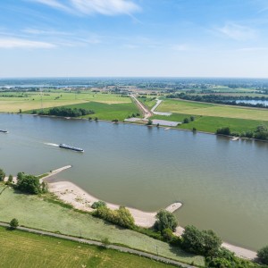 Gewässer und Umgebung: Der breite Rhein fließt horizontal durch das Bild. Die Ufer sind von grünen landwirtschaftlichen Flächen, Wiesen und vereinzelten Bäumen gesäumt. Es sind mehrere kleine Sandstrände oder Buhnen sichtbar.
Schifffahrt: Auf dem Fluss fahren zwei längliche Lastkähne (Schubverbände) in dieselbe Richtung flussaufwärts.
Baustelle: Am rechten Ufer verläuft eine aktive Baustelle diagonal durch die Felder. Es ist eine planierte, braune Schneise im Boden erkennbar, die auf Erdarbeiten für die Erdkabeltrasse hindeutet. Entlang der Trasse sind weiße Flächen, vermutlich Abdeckvlies oder Platten, zu sehen.
Hintergrund: Die Landschaft im Hintergrund ist flach und weitläufig, mit weiteren Feldern, kleinen Wäldern, Windrädern und Strommasten, die die Energieinfrastruktur der Region betonen.