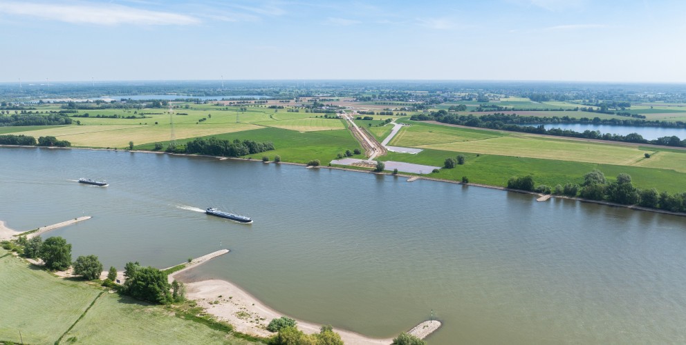 Gewässer und Umgebung: Der breite Rhein fließt horizontal durch das Bild. Die Ufer sind von grünen landwirtschaftlichen Flächen, Wiesen und vereinzelten Bäumen gesäumt. Es sind mehrere kleine Sandstrände oder Buhnen sichtbar.
Schifffahrt: Auf dem Fluss fahren zwei längliche Lastkähne (Schubverbände) in dieselbe Richtung flussaufwärts.
Baustelle: Am rechten Ufer verläuft eine aktive Baustelle diagonal durch die Felder. Es ist eine planierte, braune Schneise im Boden erkennbar, die auf Erdarbeiten für die Erdkabeltrasse hindeutet. Entlang der Trasse sind weiße Flächen, vermutlich Abdeckvlies oder Platten, zu sehen.
Hintergrund: Die Landschaft im Hintergrund ist flach und weitläufig, mit weiteren Feldern, kleinen Wäldern, Windrädern und Strommasten, die die Energieinfrastruktur der Region betonen.