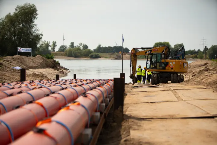 Vordergrund und Ausrüstung: Im Vordergrund liegt eine massive, aus rotem Kunststoff bestehende Rohrleitung, die durch orange Auftriebskörper zusammengehalten wird und ins Wasser ragt. Eine vertikale Metallstange mit einer kleinen Flagge markiert die Position im Wasser. Der Uferbereich besteht aus aufgeschüttetem Sand und Kies.
Hintergrund: Hinter der Baustelle erstreckt sich eine grüne Landschaft mit Bäumen und Büschen am Horizont. Weiße Banner, auf denen unter anderem "AMPRION" und "HÜLSKENS" zu lesen sind, sind am oberen Rand der Baustelle angebracht. Der Himmel ist bewölkt. Ein gelber Mobilbagger der Marke CAT ist rechts neben der Rohleitung zu sehen. 