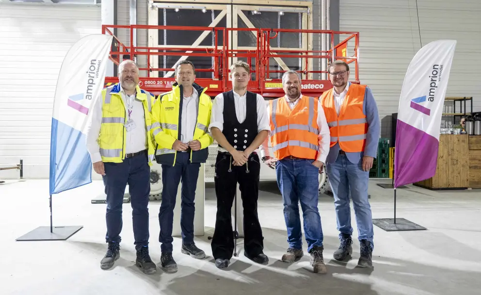 Die Gruppe besteht aus fünf Männern in unterschiedlicher Arbeits- und Geschäftskleidung. Drei Männer tragen Warnwesten in Gelb oder Orange über Hemden und Jeans. Der zweite Mann von links trägt zusätzlich zur Weste einen weißen Schutzhelm mit dem Amprion-Logo. Der Mann in der Mitte trägt traditionelle schwarze Hose mit Schlag und eine weiße Weste (Zimmererkluft), der Mann ganz rechts ein Sakko.