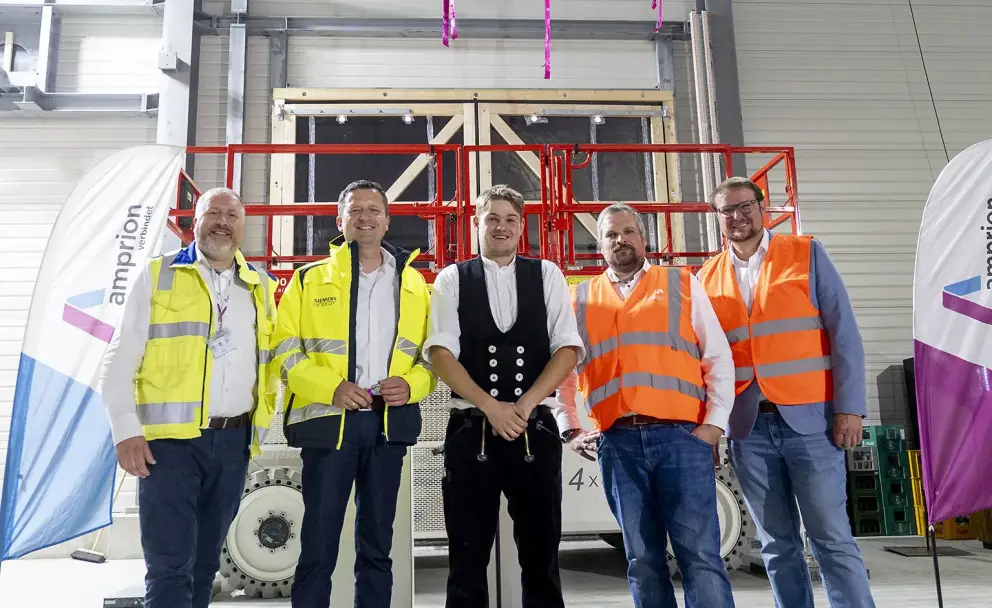 Personen: Eine Gruppe von fünf Männern, die in die Kamera blicken.
Kleidung: Zwei Männer tragen orangefarbene Warnwesten, zwei tragen gelbe Warnwesten, und ein Mann trägt eine traditionelle schwarze Hose mit Schlag und eine weiße Weste.
Ausrüstung: Der zweite Mann von links trägt einen weißen Schutzhelm.
Dekoration: Über der Gruppe hängt ein Richtkranz mit bunten Bändern.
Hintergrund: Im Hintergrund steht eine rote Scherenbühne. Links und rechts von der Gruppe stehen Banner mit dem Logo "amprion" und dem Slogan "verbindet". Die Telefonnummer "0800 20 11" ist auf einem Schild an der Scherenbühne sichtbar. An der Wand steht der Text "4x".