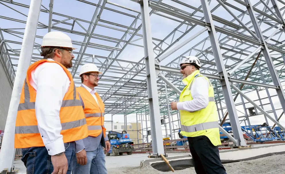 Personen: Drei Männer sind in der Szene zu sehen.
Der Mann links trägt einen weißen Schutzhelm, eine orangefarbene Warnweste über einem weißen Langarmhemd, Jeans und hat einen dunklen Bart.
Der Mann in der Mitte trägt ebenfalls einen weißen Schutzhelm und eine orangefarbene Warnweste, jedoch über einem grauen Sakko und einem weißen Hemd, kombiniert mit blauen Jeans.
Der Mann rechts trägt einen weißen Schutzhelm, eine gelbe Warnweste über einem weißen Hemd, schwarze Hosen und eine Brille. Er steht den anderen beiden zugewandt und gestikuliert.