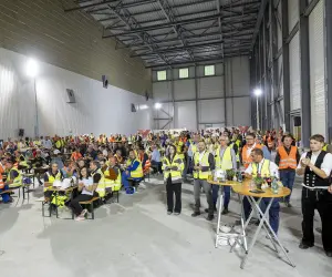 Ort: Die Szene spielt sich im Inneren einer im Bau befindlichen Industriehalle ab, erkennbar an den hellen Trapezblechwänden und der sichtbaren Deckenkonstruktion [1.1].
Personen und Kleidung: Rund 200 Bauarbeiter und Projektmitarbeiter sitzen an langen Holztischen oder stehen im Vordergrund. Viele tragen orangefarbene oder gelbe Warnwesten über ihrer Kleidung, einige tragen Schutzhelme auf den Tischen 