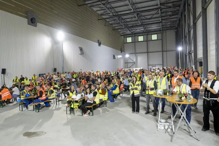 Ort: Die Szene spielt sich im Inneren einer im Bau befindlichen Industriehalle ab, erkennbar an den hellen Trapezblechwänden und der sichtbaren Deckenkonstruktion [1.1].
Personen und Kleidung: Rund 200 Bauarbeiter und Projektmitarbeiter sitzen an langen Holztischen oder stehen im Vordergrund. Viele tragen orangefarbene oder gelbe Warnwesten über ihrer Kleidung, einige tragen Schutzhelme auf den Tischen 