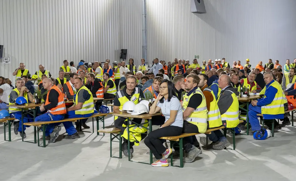 Zahlreiche Personen, darunter viele Bauarbeiter und Projektmitarbeiter in orangefarbenen und gelben Warnwesten, sitzen an langen Holztischen in einer großen, industriellen Halle.