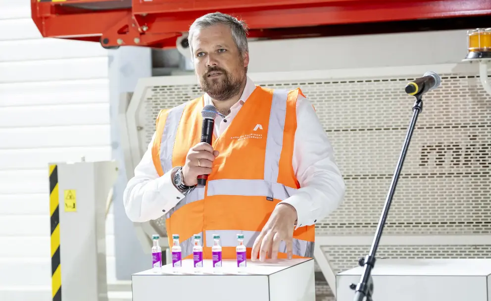 Person: Ein bärtiger Mann mittleren Alters trägt eine orangefarbene Warnweste mit reflektierenden Streifen über einem weißen, langärmeligen Hemd und einer Armbanduhr. Auf der Brust der Weste ist ein kleines, weißes Logo erkennbar.
Vordergrund: Auf einem weißen Podest vor dem Mann stehen sechs kleine, transparente Fläschchen mit pinkfarbener Flüssigkeit. Rechts im Bild befindet sich ein Mikrofonständer.
Hintergrund: Im Hintergrund ist eine große, rot lackierte Metallkonstruktion mit gelben und schwarzen Warnmarkierungen und weißem Text, der unter anderem "0800 freecall" anzeigt, zu sehen. Links davon ist eine gelb-schwarz markierte vertikale Säule. 
