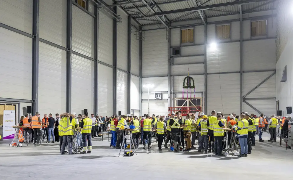 Zahlreiche Personen, darunter viele Bauarbeiter und Projektmitarbeiter in orangefarbenen und gelben Warnwesten, stehen oder sitzen an langen Holztischen in einer großen, industriellen Halle.