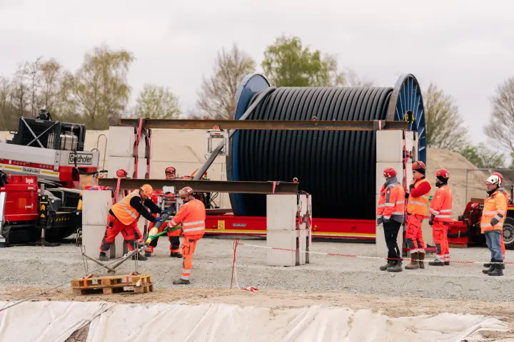 Aktivitäten und Personen: Im Zentrum des Bildes sind mehrere Bauarbeiter in orangefarbener Warnschutzkleidung und Schutzhelmen mit der Handhabung der Trommel beschäftigt. Zwei Arbeiter im linken Vordergrund verwenden Werkzeuge, um Befestigungen zu lösen oder anzubringen.
Ausrüstung: Eine massive, schwarze Kabeltrommel mit blauen Flanschen ruht auf einer roten Transportvorrichtung, die wiederum auf großen weißen Betonblöcken platziert ist. Links im Bild ist der Ausleger eines roten Minikrans oder Baggers mit dem Schriftzug "Goldhofer" und "Wiedhofer" zu sehen. Auf der rechten Seite der Szene stehen weitere Bauarbeiter und der hintere Teil eines Fahrzeugs.
Baustellenbereich: Die Szene spielt sich auf einer Baustelle mit geschottertem Boden ab. Im Vordergrund liegt eine Palette, und der Boden ist teilweise mit weißem Vlies bedeckt. Im Hintergrund begrenzen graue Wände das Gelände, und dahinter sind Bäume unter einem bewölkten Himmel sichtbar.