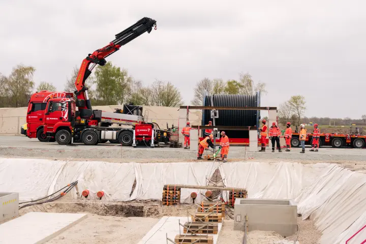 Aktivitäten: Im Zentrum des Bildes entlädt ein roter LKW-Ladekran der Marke Palfinger eine große, schwarze Kabeltrommel von einem Tieflader. Mehrere Bauarbeiter in orangefarbener Schutzkleidung und Schutzhelmen sind beteiligt, um den Vorgang zu sichern und zu koordinieren.
Ausrüstung: Die Kabeltrommel hat massive Ausmaße und wiegt typischerweise zwischen 50 und 90 Tonnen. Der Kran ist auf einem roten MAN-LKW montiert, der an der Seite unter anderem den Schriftzug "DE ROMEIN" trägt. Neben dem LKW steht der Tieflader.
Baustellenbereich: Die Szene spielt sich an einer großen Baugrube ab, deren Böschungen mit weißem Vlies ausgekleidet sind, um den Boden zu schützen. Im Vordergrund sind vorbereitete Betonfundamente und Holzpaletten erkennbar. Im Hintergrund begrenzen graue Betonwände und dahinter Bäume die Baustelle. 