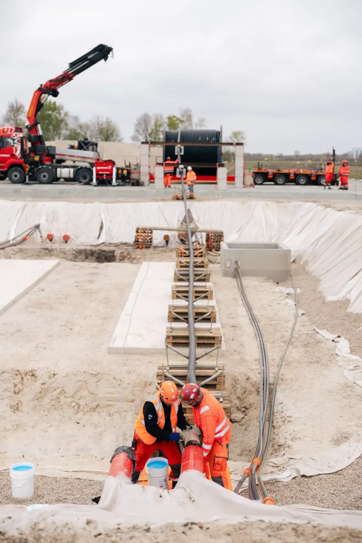 Aktivitäten und Personen: Im Zentrum des Bildes entlädt ein roter LKW-Ladekran der Marke Palfinger eine große, schwarze Kabeltrommel von einem Tieflader. Mehrere Bauarbeiter in orangefarbener Schutzkleidung und Schutzhelmen sind beteiligt, um den Vorgang zu sichern und zu koordinieren.
Ausrüstung: Die Kabeltrommel hat massive Ausmaße und wiegt typischerweise zwischen 50 und 90 Tonnen. Der Kran ist auf einem roten MAN-LKW montiert, der an der Seite unter anderem den Schriftzug "DE ROMEIN" trägt.
Baustellenbereich: Die Szene spielt sich an einer großen Baugrube ab, deren Böschungen mit weißem Vlies ausgekleidet sind, um den Boden zu schützen. Im Vordergrund sind vorbereitete Betonfundamente und Holzpaletten erkennbar. Im Hintergrund begrenzen graue Betonwände und dahinter Bäume die Baustelle. 