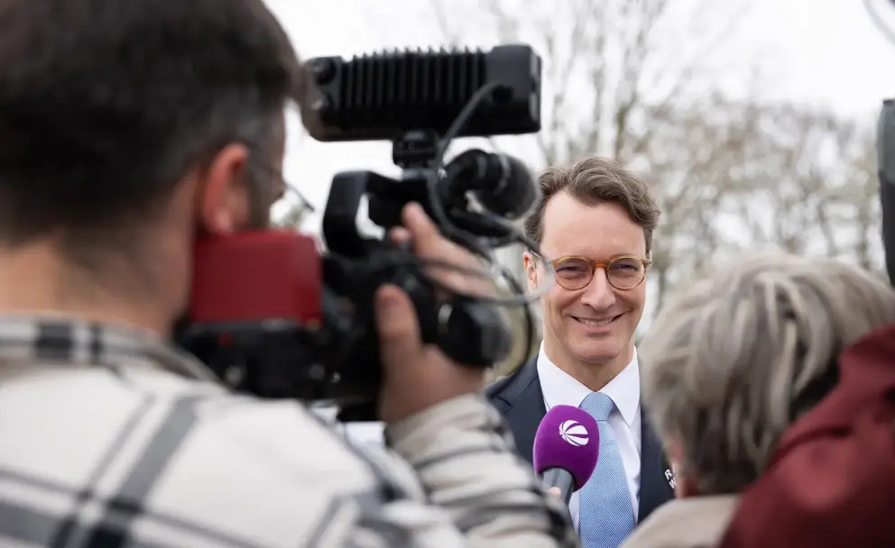 Das Bild zeigt eine Nahaufnahme eines Mannes mit hellbraunen, leicht gewellten Haaren und Brille, der in die Kamera lächelt. Er trägt einen dunklen Anzug, ein weißes Hemd und eine hellblaue Krawatte mit einem dezenten Muster. Vor seinem Mund ist ein lilafarbenes Mikrofon mit einem weißen Logo platziert, das wie ein stilisiertes "e" aussieht. Das Mikrofon wird von einer Person gehalten, die außerhalb des Bildrahmens zu sein scheint.
Im Vordergrund links ist der Rücken einer Person mit dunklen, kurzen Haaren zu sehen, die eine Videokamera bedient. Die Kamera ist schwarz mit roten Akzenten und einem großen Objektiv. Ein Mikrofon mit einem Windschutz ist oben an der Kamera befestigt und ragt in den oberen Bildrand.
Rechts im Vordergrund ist der Hinterkopf einer Person mit kurzen, grauen Haaren zu sehen, die dem Mann mit dem Mikrofon zuzuhören scheint.
Der Hintergrund ist unscharf und zeigt helle, möglicherweise bewölkte oder neblige Bedingungen mit verschwommenen Bäumen oder Strukturen. 