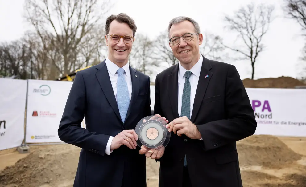Das Bild zeigt zwei Männer in Anzügen, die nebeneinander stehen und in die Kamera lächeln. Der Mann links hat braune, leicht gewellte Haare und trägt eine Brille, einen dunkelblauen Anzug, ein weißes Hemd und eine hellblaue Krawatte mit weißem Muster. Er hält mit beiden Händen eine transparente, runde Plakette mit einem kupferfarbenen Zentrum und konzentrischen Kreisen.
Der Mann rechts hat graue, kurze Haare und trägt ebenfalls eine Brille, einen schwarzen Anzug, ein weißes Hemd und eine grüne Krawatte mit einem Muster. Er hält die Plakette ebenfalls mit beiden Händen und blickt freundlich in die Kamera.
Im Hintergrund sind unscharf Teile einer Baustelle zu erkennen. Es gibt einen sandigen oder erdfarbenen Boden, einen weißen Bauzaun mit Aufschriften und Logos (teilweise lesbar ist "A-Nord") und einige kahle Bäume. Der Himmel ist grau und bedeckt, was auf einen trüben Tag hindeutet.