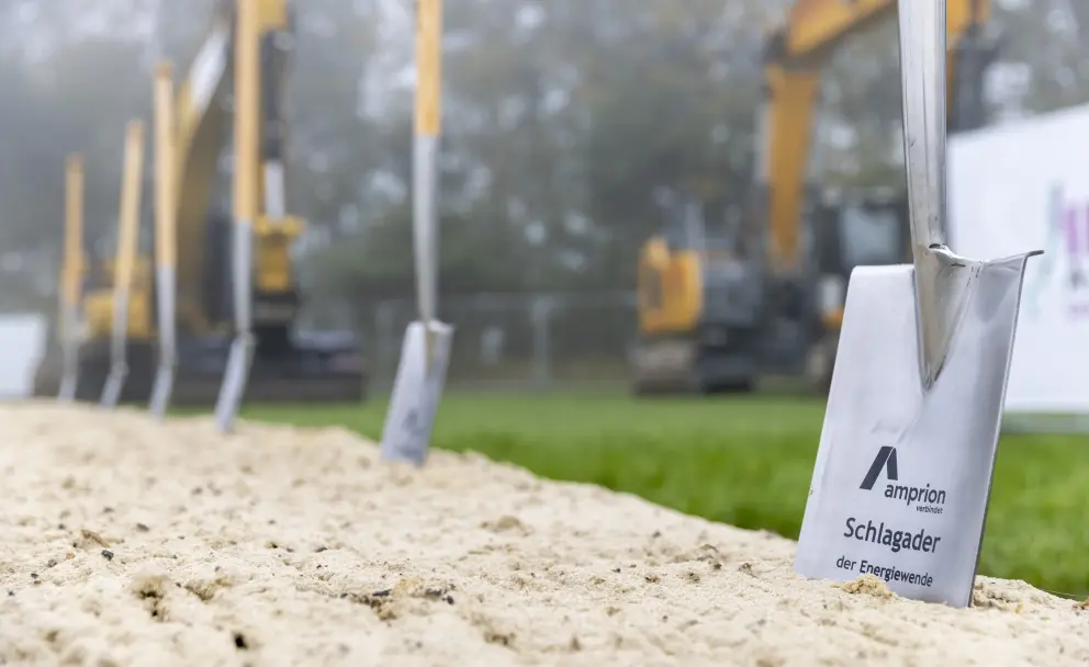 Symbolische Spaten: Im Vordergrund sind mehrere Spaten mit Holzstielen und silbernen Blättern in den Sand gesteckt. Einer der vordersten Spaten trägt ein Schild mit dem Logo und dem Slogan des Übertragungsnetzbetreibers Amprion, auf dem "Amprion verbindet" und "Schlagader der Energiewende" zu lesen ist.
Hintergrund: Im unscharfen Hintergrund, eingehüllt in Nebel oder Dunst, sind gelbe Baumaschinen (Bagger) erkennbar. Rechts ist ein weißes Banner oder Schild aufgestellt.