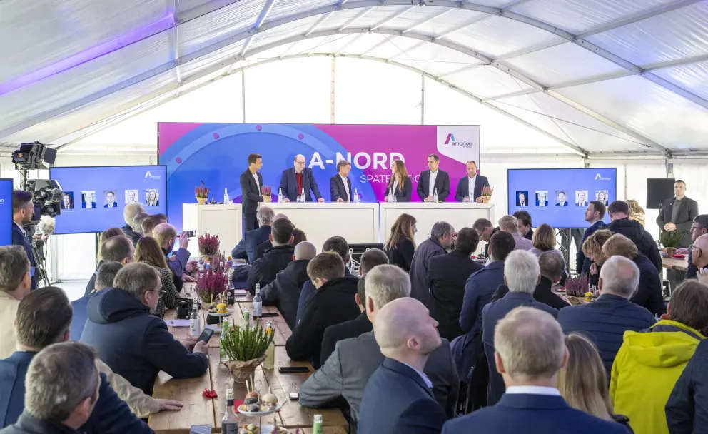 Das Foto zeigt eine Podiumsdiskussion im Inneren eines großen weißen Veranstaltungszeltes, in dem zahlreiche Personen an Holztischen Platz genommen haben. Die Szene ist von oben herab aufgenommen und fängt die Atmosphäre der Veranstaltung ein.