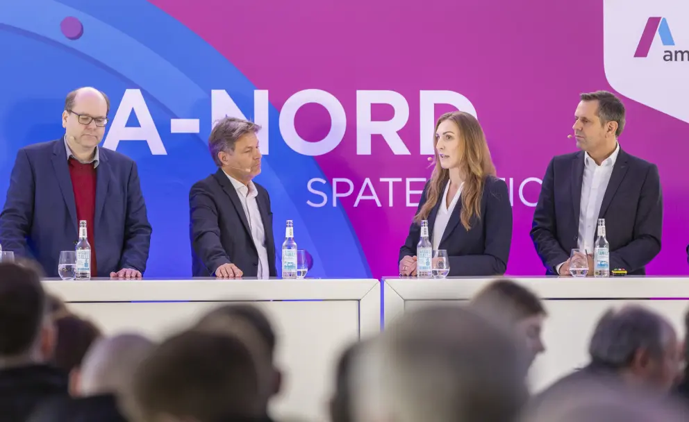 Die Teilnehmer am Podium sind von links nach rechts:
Erste Person (links außen): Ein Mann mit Brille und grauem Haar, der einen dunkelblauen Anzug über einem roten Pullover trägt.
Zweite Person (links mitte): Robert Habeck, der deutsche Bundesminister für Wirtschaft und Klimaschutz. Er trägt einen dunkelblauen Anzug mit einem weißen Hemd und blickt nach rechts zur dritten Person.
Dritte Person (rechts mitte): Eine Frau mit langen braunen Haaren, die einen dunkelblauen Hosenanzug mit einer weißen Bluse trägt. Sie spricht gerade in die Richtung der vierten Person und des Publikums.
Vierte Person (rechts außen): Ein Mann mit kurzem, dunklem Haar, der einen dunkelblauen Anzug mit einem weißen Hemd trägt und nach vorne blickt. 
