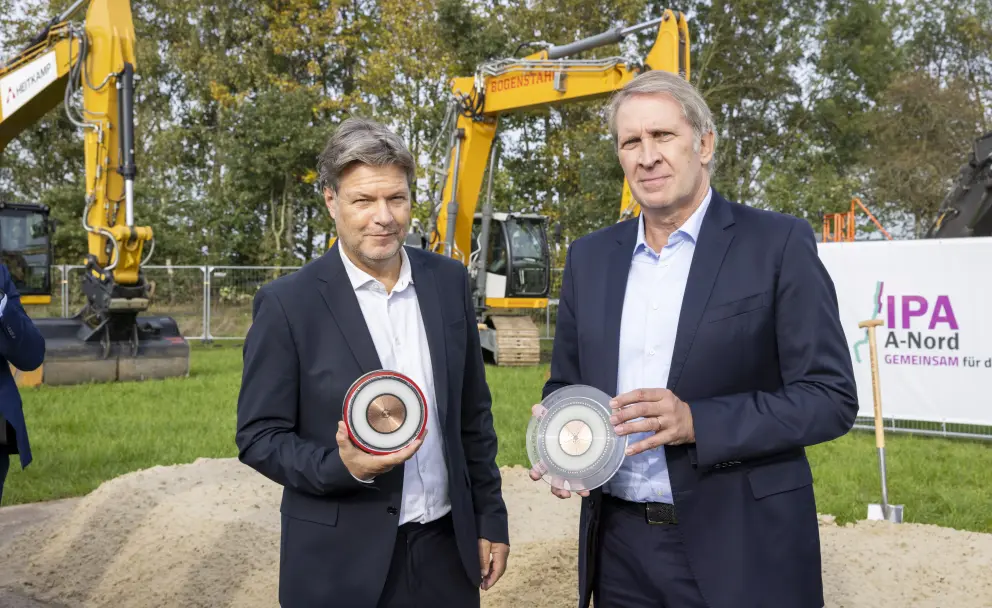 Das Foto zeigt eine Szene im Freien, aufgenommen während des offiziellen Spatenstichs für die Erdkabel-Stromtrasse A-Nord. Im Vordergrund, auf einer Rasenfläche stehend und vor einem kleinen Sandhaufen, sind zwei Männer in Anzügen zu sehen, die in die Kamera blicken:
Links steht Robert Habeck, der deutsche Bundesminister für Wirtschaft und Klimaschutz. Er trägt einen dunkelblauen Anzug über einem hellblauen Hemd und hält in seinen Händen ein kreisförmiges, rot ummanteltes Stromkabelmuster.
Rechts steht Dr. Hans-Jürgen Brick, der damalige Vorsitzende der Geschäftsführung (CEO) des Übertragungsnetzbetreibers Amprion. Auch er trägt einen dunkelblauen Anzug über einem hellblauen Hemd und hält ein identisches, aber transparentes Kabelmuster in den Händen.