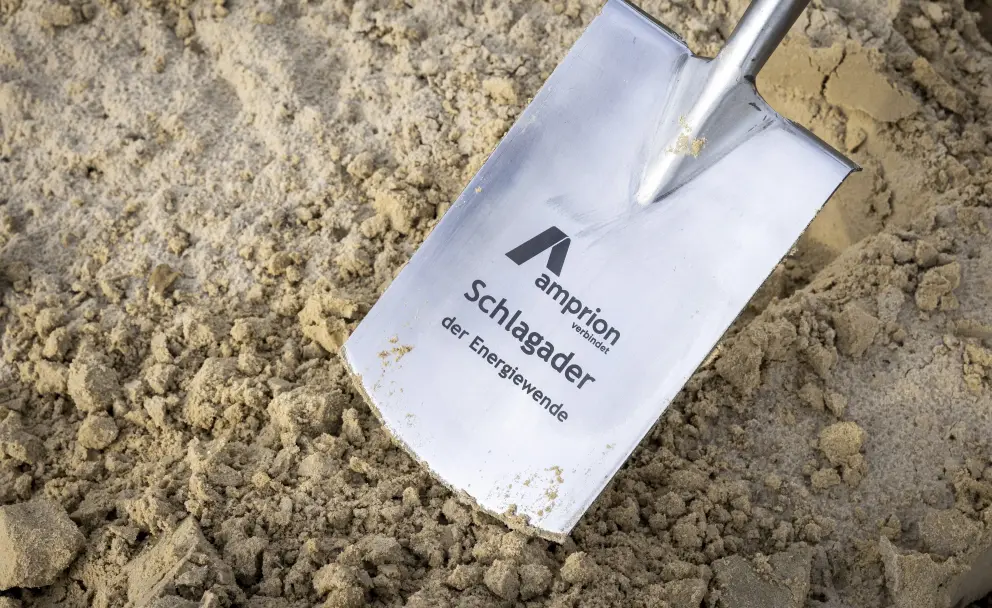 Nahaufnahme der Schaufel eines Spatens, der in sandiger Erde steckt. Auf dem metallischen Schaufelblatt ist das schwarze Logo der Firma Amprion abgebildet, gefolgt vom Slogan "amprion verbindet" sowie in größeren Buchstaben "Schlagader der Energiewende". Der Hintergrund besteht nur aus unscharfem Sand.