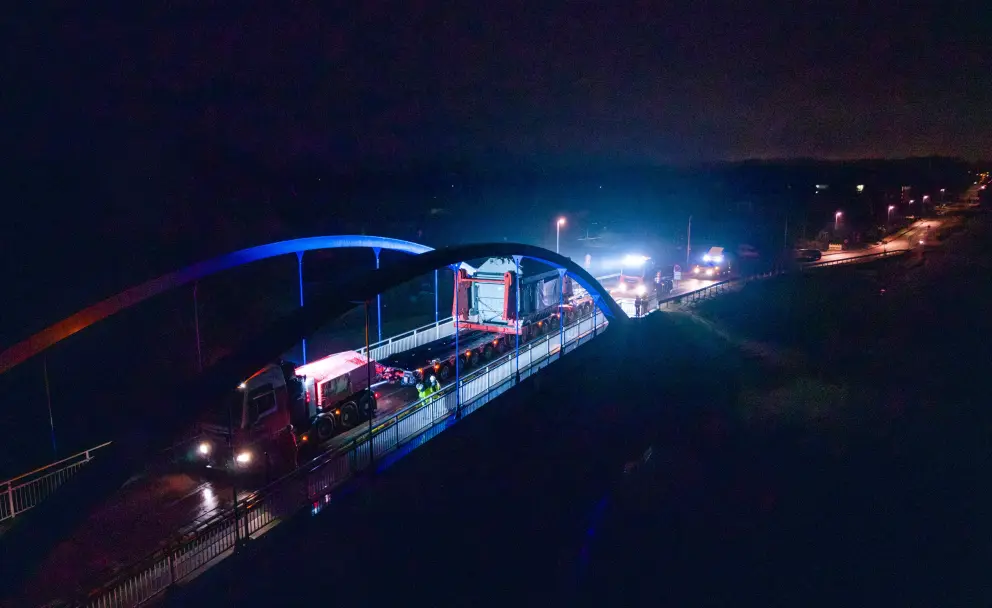 Ein großer Transformator wird nachts auf einem Selbstfahrer-Modultransporter (SPMT) über eine blaue Bogenbrücke gefahren.