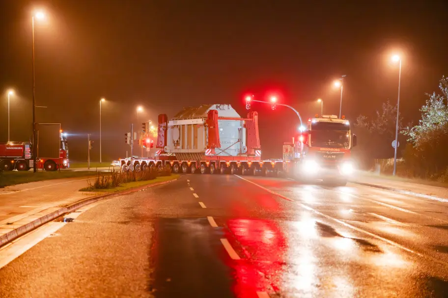 Ein großer Transformator wird nachts auf einem Selbstfahrer-Modultransporter (SPMT) über eine Straße transportiert. Warnleuchten leuchten rot.