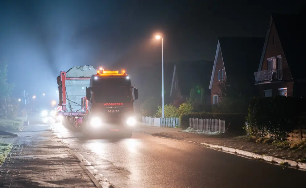 Ein Schwertransporter fährt nachts mit einem Transformator auf einem nassen Industrieweg.