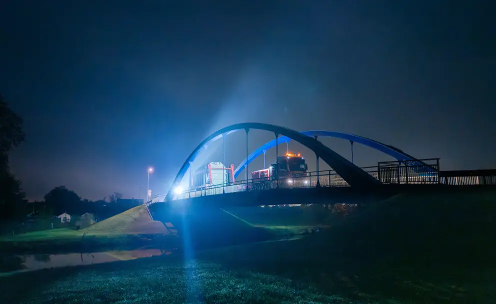 Ein großer Schwertransport mit einem Transformator fährt nachts über eine blau beleuchtete Bogenbrücke.