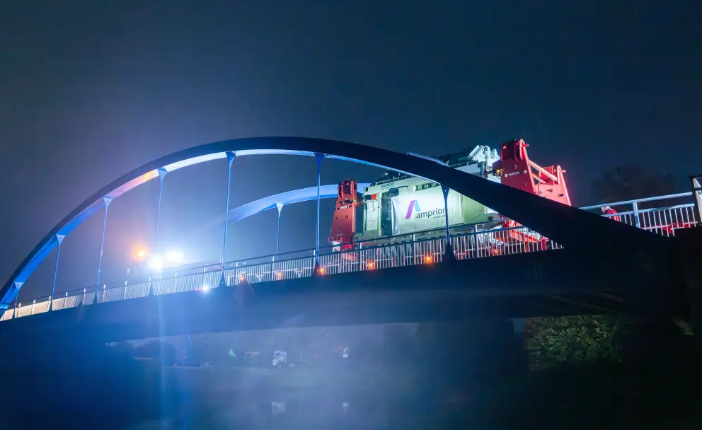 Ein großer Schwertransport mit einem Transformator fährt nachts über eine blau beleuchtete Bogenbrücke.