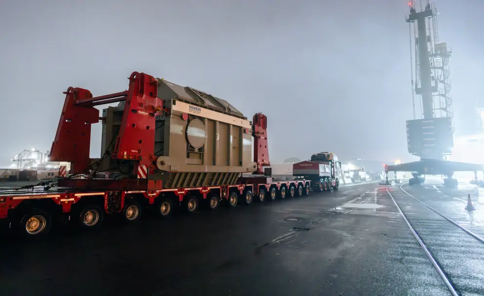 Ein großer Transformator wird nachts auf einem Selbstfahrer-Modultransporter in einem Industriehafen entladen.