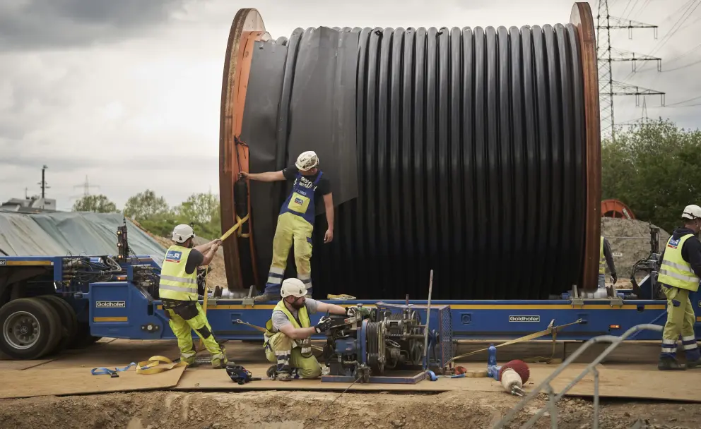 
Auf dem Bild ist eine Baustelle zu sehen, auf der mehrere Arbeiter damit beschäftigt sind, ein riesiges Kabel von einer großen Trommel abzuwickeln. Die Trommel ist auf einem blauen Tieflader-Anhänger montiert.