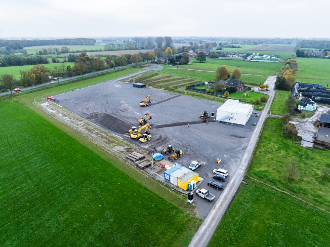 Das Bild zeigt eine Luftaufnahme einer aktiven Baustelle in einer ländlichen Umgebung, umgeben von grünen Feldern und Wiesen.
Beschreibung des Bildes
Das zentrale Element ist eine große, rechteckige, grau-braune Schotterfläche. Auf dieser Baustellenfläche befinden sich mehrere Baumaschinen, darunter große gelbe Bagger, sowie kleinere Fahrzeuge wie Lieferwagen und PKWs. Im unteren Bereich der Fläche steht ein blauer Container und eine weiße Zeltstruktur im mittleren rechten Bereich.
Die Baustelle ist klar abgegrenzt von der umliegenden Landschaft. Die Umgebung ist geprägt von weitläufigen grünen Wiesen und Feldern. Im oberen Teil des Bildes verläuft eine Straße, gesäumt von Bäumen, und weiter hinten sind einige Gebäude und Höfe erkennbar. Der Himmel ist bedeckt und grau.
