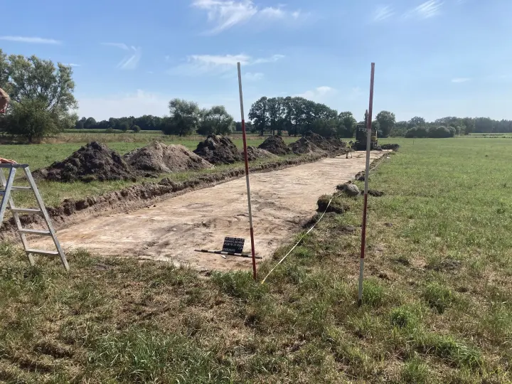 Archäologische Ausgrabung in einem länglichen Graben auf einer Wiese, mit Erdhaufen und Vermessungspfählen.