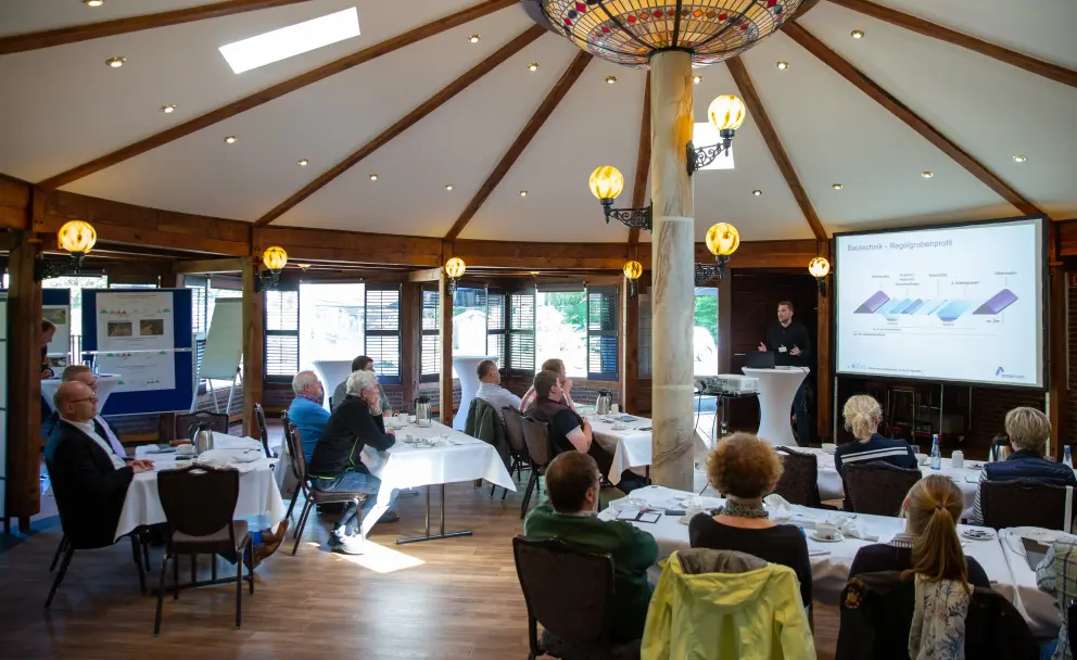 Die Szene zeigt eine Informationsveranstaltung in einem achteckigen Raum mit Holzbalkendecke und orange getönten Hängelampen. Mehrere Personen sitzen an weiß gedeckten Tischen und blicken nach vorne. Links im Bild sind zusätzliche Informationstafeln auf einem Ständer zu sehen. Zentral steht ein Mann am Rednerpult und gestikuliert in Richtung der Leinwand. Auf der Leinwand ist eine Präsentationsfolie mit der Aufschrift "Bauwirtsch. & Regelprüfverf." und einer schematischen Darstellung verschiedener Bodenschichten in blau und lila zu sehen.