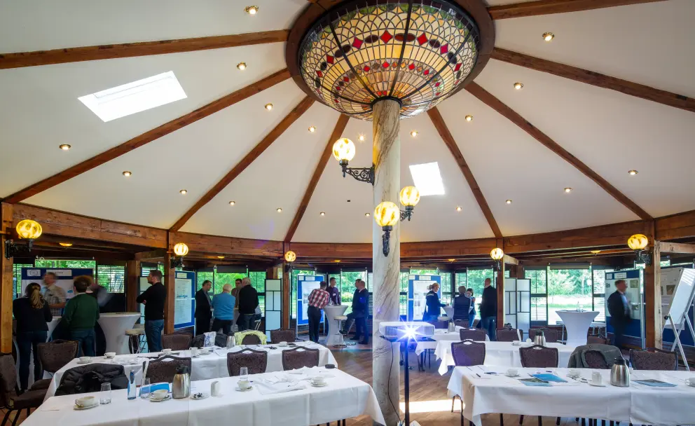 Das Bild zeigt einen großen, runden Tagungsraum mit einer hohen, zeltförmigen Decke, die von Holzbalken getragen wird. In der Mitte des Raumes steht ein dicker, baumstammähnlicher Pfeiler, an dem eine große, verzierte Glasleuchte im Tiffany-Stil und mehrere kleinere, gelbe Kugelleuchten befestigt sind.
Der Raum ist für eine Veranstaltung vorbereitet. Im Vordergrund stehen mehrere Tische, die mit weißen Tischdecken, Besteck und Kaffeekannen gedeckt sind. Im mittleren Bereich sind diverse Personen in Gruppen stehend zu sehen, die sich unterhalten. Dort befinden sich auch Stehtische und mehrere weiße Informationstafeln mit blauen Rändern.
Der Raum ist von großen Fenstern umgeben, die den Blick auf viel Grün und Bäume freigeben. Mehrere kleine Deckenstrahler sorgen für zusätzliche Beleuchtung.