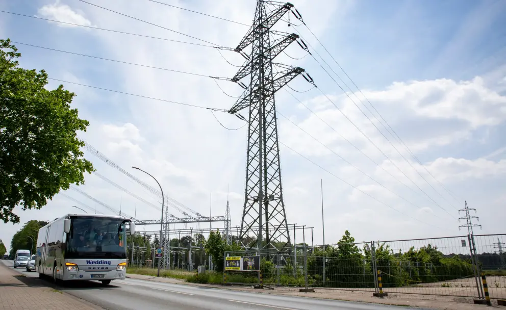 Die Fotografie zeigt eine Straßenszene an einem sonnigen Tag. Links auf der Fahrbahn fährt ein weißer Reisebus, auf dessen digitaler Anzeige vorne "Wedding" steht. Die Straße führt an einer eingezäunten Industrieanlage vorbei. In der Mitte des Bildes dominiert ein hoher, metallener Freileitungsmast (Gittermast) mit zahlreichen Isolatoren und gespannten Hochspannungsleitungen, die sich über das Bild erstrecken. Im Hintergrund sind das Gelände einer Umspannstation mit weiteren Masten und Strukturen sowie blauer Himmel mit weißen Wolken zu sehen. Rechts verläuft ein Bauzaun.