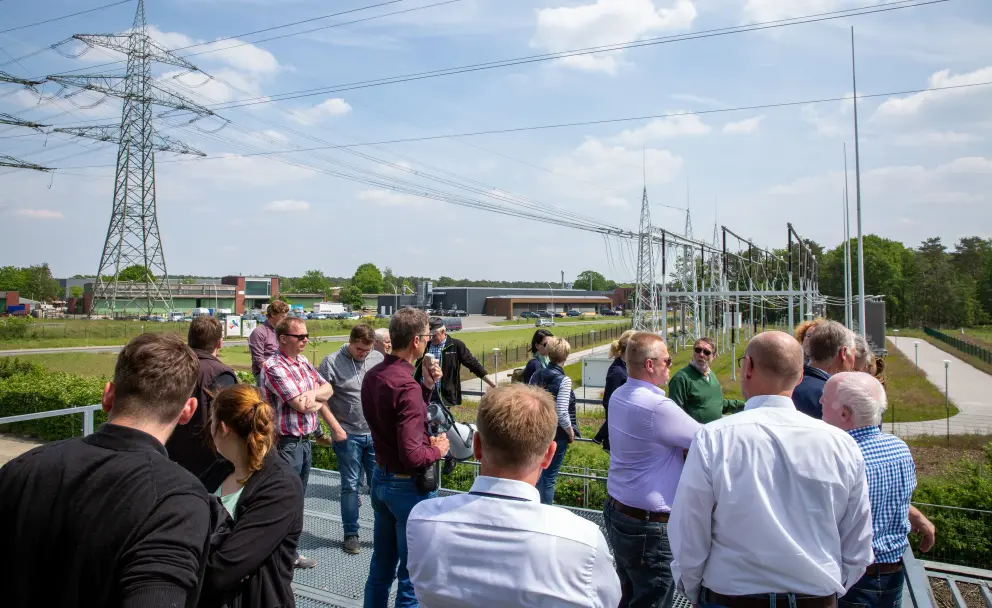Eine Gruppe von Menschen auf einer erhöhten Plattform betrachtet eine Umspannstation und Hochspannungsmasten.