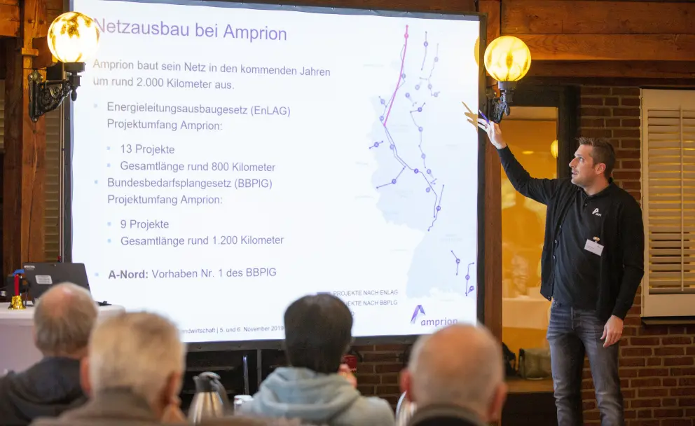Eine Innenaufnahme zeigt einen Mann mittleren Alters in einem schwarzen Poloshirt und einer schwarzen Strickjacke, der einen Vortrag hält. Er steht vor einer Projektionsfläche, auf der eine Präsentation über den "Netzausbau bei Amprion" angezeigt wird. Der Redner hält mit seiner rechten Hand einen Pointer und deutet auf eine Karte von Deutschland, auf der geplante Stromtrassen und Standorte markiert sind. Die Folie listet Details zu 13 EnLAG-Projekten (rund 800 km) und 9 BBPIG-Projekten (rund 1.200 km) auf, einschließlich des A-Nord-Projekts. Auf einem Tisch im Vordergrund stehen ein Laptop, eine Glasflasche Wasser und ein Weinglas. Im Hintergrund sind dunkle Holzvertäfelungen, Ziegelmauern und zwei dekorative, beleuchtete Wandleuchten mit orangefarbenen Schirmen zu sehen. Die Zuhörer im Vordergrund, erkennbar an ihren Hinterköpfen, verfolgen den Vortrag.
