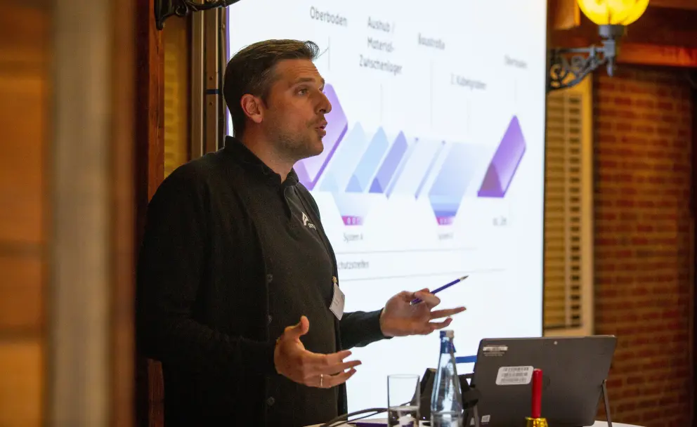 Eine Innenaufnahme zeigt einen Mann mittleren Alters in einem schwarzen Poloshirt und einer schwarzen Strickjacke, der einen Vortrag hält. Er steht vor einer Projektionsfläche, auf der eine technische Infografik angezeigt wird, die verschiedene Phasen eines Prozesses darstellt: "Oberboden", "Aushub / Material / Zwischenlager", "Baustoffe". Der Mann hält einen blauen Stift in der rechten Hand und gestikuliert mit beiden Händen, während er zu einem unsichtbaren Publikum spricht. Auf einem Tisch vor ihm stehen ein Laptop, eine Glasflasche Wasser und ein Weinglas. Im Hintergrund sind dunkle Holzvertäfelungen, eine verzierte schwarze Wandhalterung und eine gelbe Wandleuchte zu sehen, die eine warme, gedämpfte Beleuchtung erzeugt.
