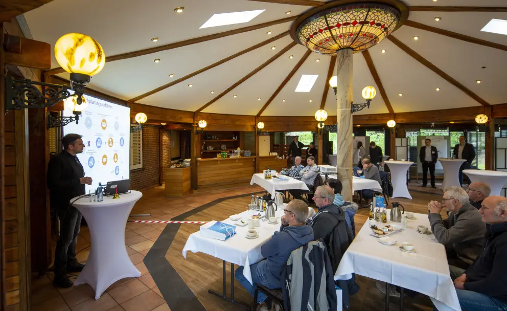 Eine Innenaufnahme aus der Perspektive eines Zuschauers in einem Konferenzraum mit einer sechseckigen Holzdecke und einer großen, runden Deckenleuchte im Tiffany-Stil. Im linken Vordergrund steht ein Redner mit einem schwarzen Oberteil und Jeans vor einem weißen Stehtisch und einem Flipchart, das eine technische Infografik zeigt. Die Grafik enthält kleine Diagramme und textliche Elemente wie "Data (current)" und "Real World Evidence". Die Zuhörer sitzen an weiß gedeckten Tischen und verfolgen den Vortrag. Im rechten Hintergrund stehen weitere Personen an Stehtischen, und durch große Fenster ist unscharfe grüne Vegetation zu sehen. 