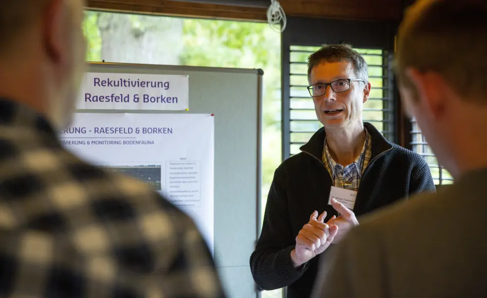 Eine Innenaufnahme aus der Vogelperspektive zeigt drei Männer, die sich in einem Holzraum um ein schwarzes Rednerpult versammelt haben. Der Mann in der Mitte trägt einen schwarzen Pullover, eine Brille und spricht, während er mit seinen Händen gestikuliert. Der Mann links im Vordergrund ist nur von hinten zu sehen und trägt ein kariertes Oberteil. Der Mann rechts im Vordergrund ist ebenfalls nur von hinten zu sehen und trägt ein graues T-Shirt. Eine weiße Präsentationswand hinter den Männern zeigt oben links in schwarzer Schrift "Rekultivierung Raesfeld & Borken". Im Hintergrund sind durch ein Fenster unscharf grüne Bäume zu sehen.
