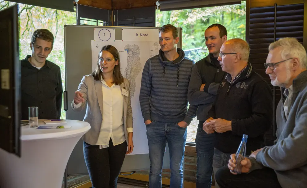 Eine Innenaufnahme zeigt eine Gruppe von sechs Erwachsenen, die sich in einem Holzraum um einen weißen Stehtisch versammelt haben. Links steht eine Frau in einem hellgrauen Blazer über einem weißen Hemd mit einer Brille und gestikuliert mit ihrer rechten Hand in Richtung eines Mannes in der Mitte, als würde sie etwas erklären. Der Mann in der Mitte, mit dunkelblondem Haar und einem gestreiften Hoodie, steht mit in den Hosentaschen vergrabenen Händen da und schaut in ihre Richtung. Hinter ihnen steht eine weiße Präsentationswand, auf der oben rechts in schwarzer Schrift "A-Nord" zu lesen ist. Auf der rechten Seite der Gruppe steht ein Mann mittleren Alters, der dem Gespräch zuhört, und ganz rechts ein älterer Mann mit Brille, der ebenfalls aufmerksam ist. Durch große Fenster im Hintergrund ist unscharf grünes Herbstlaub zu sehen.