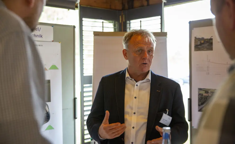 Eine frontale Nahaufnahme im Innenraum eines Vortragssaals oder Konferenzraums. Ein Mann mittleren Alters mit kurzen grauen Haaren und einem Ziegenbart trägt einen schwarzen Blazer über einem weißen Hemd mit kleinen Karomustern. Er steht vor einer weißen Präsentationswand und einem Flipchart, gestikuliert mit beiden Händen und spricht ein unsichtbares Publikum an. 