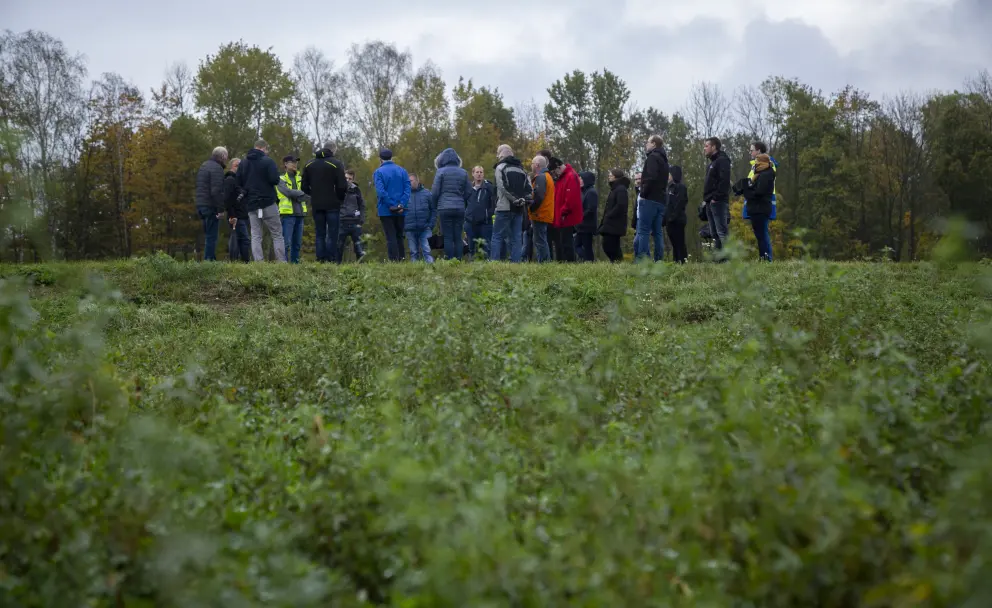 Eine Außenaufnahme zeigt eine Gruppe von etwa 15 bis 20 Personen auf einem erhöhten, grasbewachsenen Ufer oder Damm. Die Teilnehmer tragen wetterfeste Freizeitkleidung in dunklen Farbtönen, darunter Jacken in Schwarz, Blau, Grau sowie einige Farbakzente wie eine gelbe Warnweste, eine hellblaue Jacke und eine orangefarbene Jacke. Sie stehen in lockeren Gruppen und blicken nach links oder in die Mitte, wo sie sich unterhalten. Der Vordergrund des Bildes ist dominiert von unscharfer, dunkelgrüner Vegetation. Im Hintergrund erstreckt sich ein dichter Waldrand mit Laubbäumen in herbstlichen Grün- und Gelbtönen, über denen ein bewölkter, grauer Himmel zu sehen ist.
